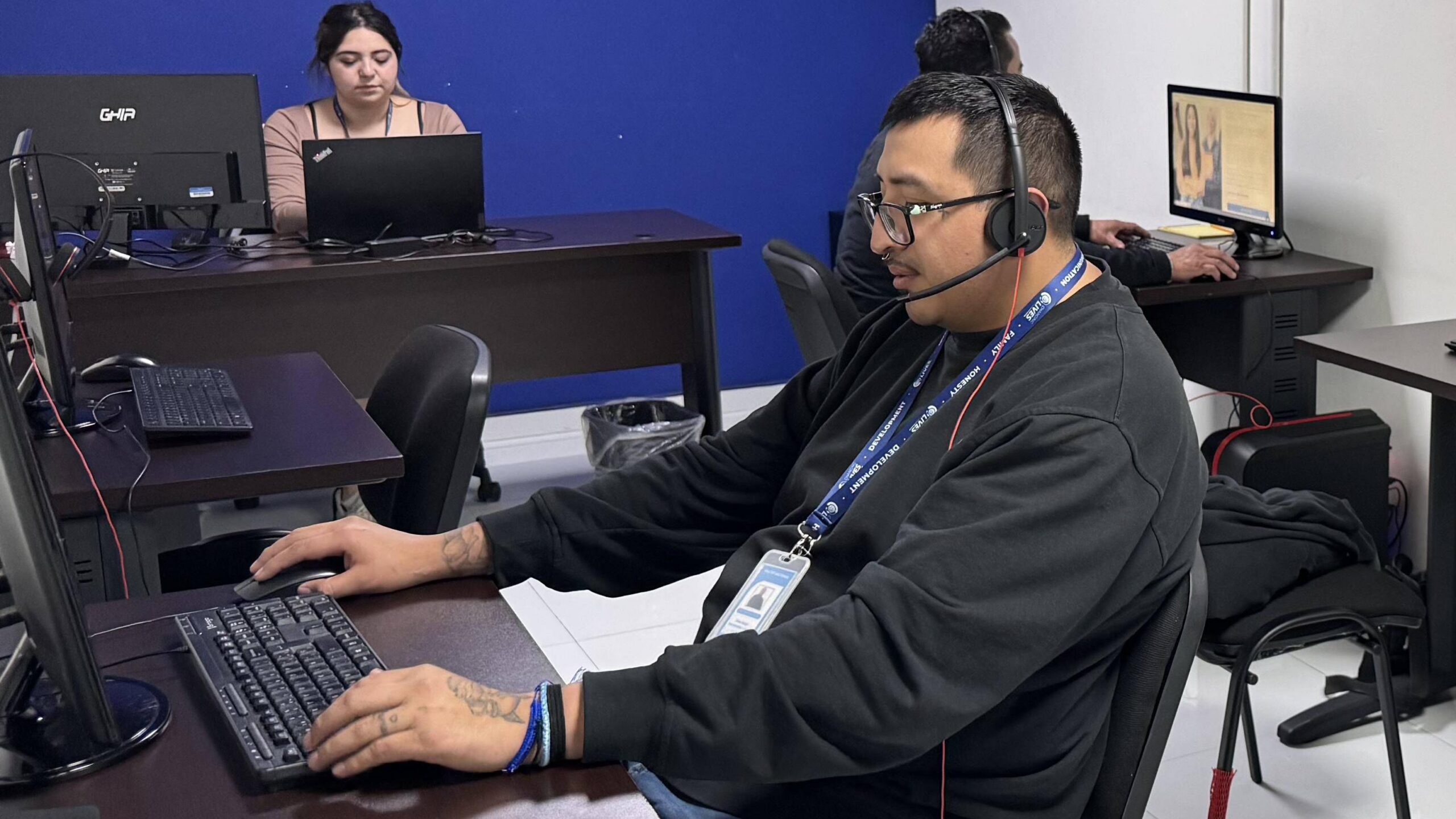 Hombre con auriculares trabajando en una computadora en una oficina compartida, con otras personas usando equipos al fondo.