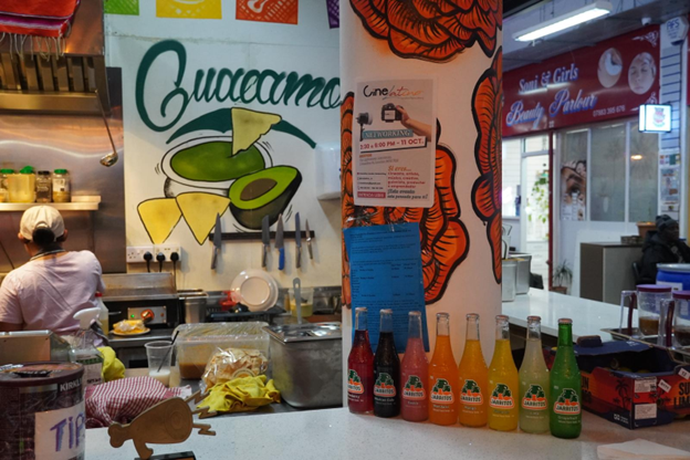 Se ve un mostrador del restaurante. y sobre este una hilera de botellas con soda. en la pared del fondo hay una ilustraci&oacute;n de un aguacate con tortillas y la palabra "guacamole". Se a una empleada de espaldas cocinando. 
