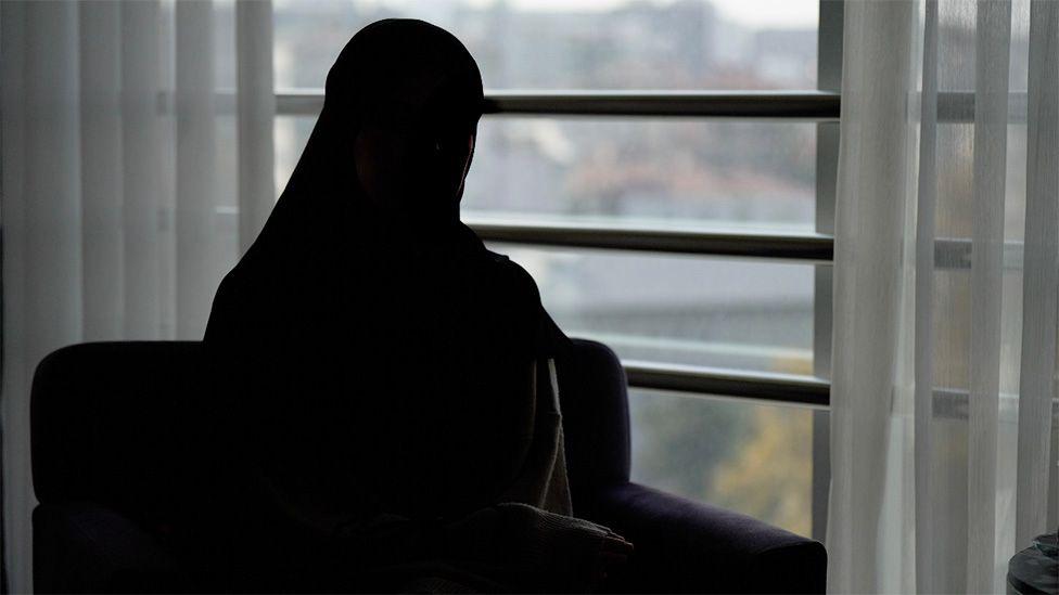 Amal aparece completamente recortada contra la luz, sentada en el interior junto a un gran ventanal con cortinas transparentes, con la mirada puesta en un paisaje urbano difuminado.