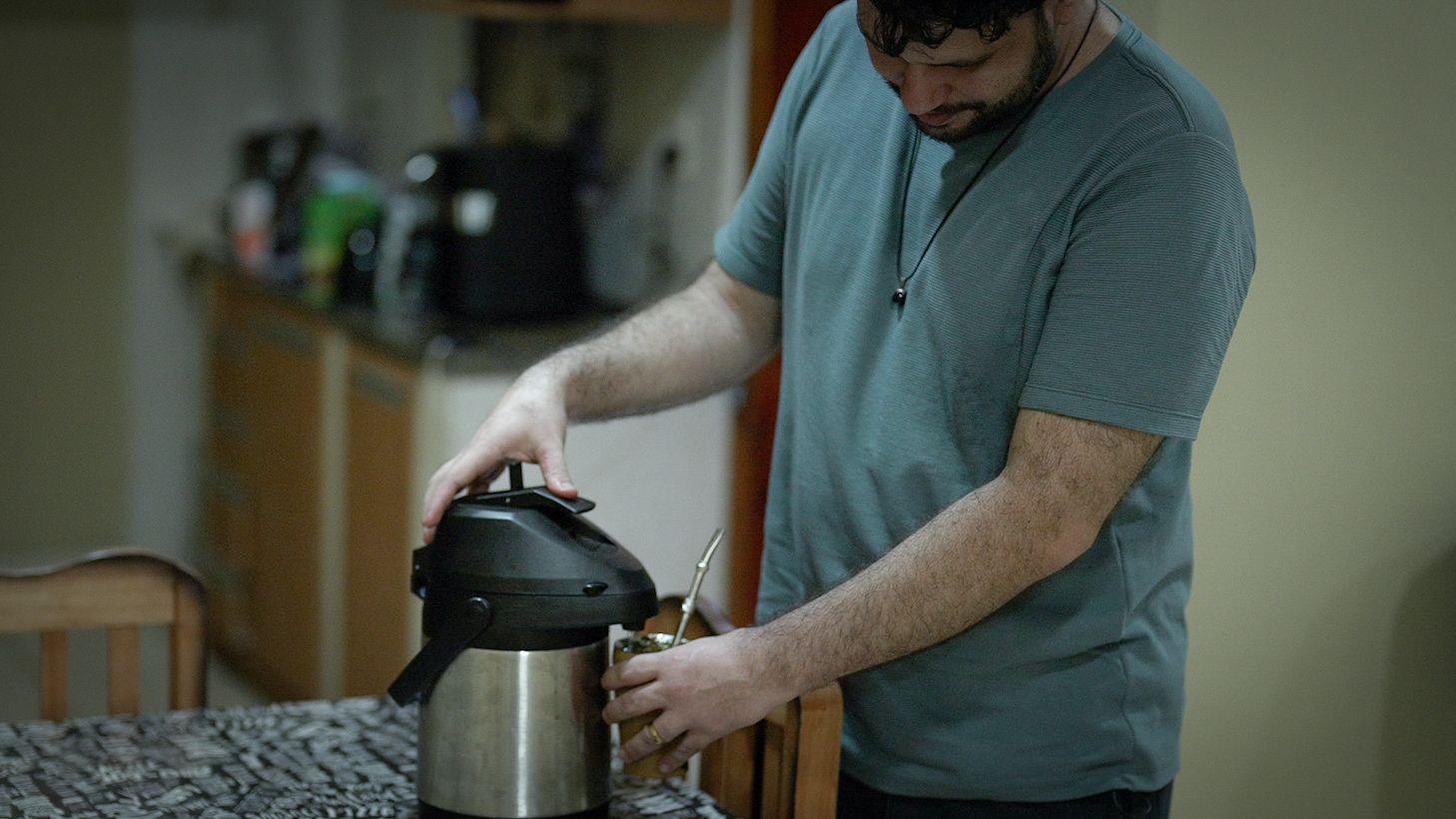 Un hombre con playera verde, prepara en una cocina su infusi&oacute;n de yerba mate con agua de un termo.