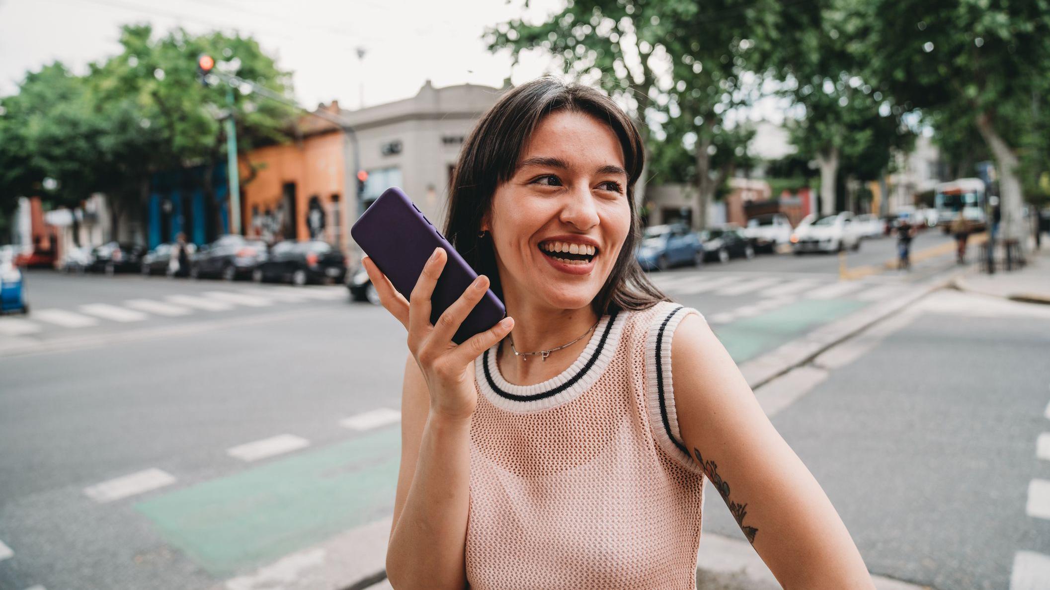una mujer en medio de una calle env&iacute;a una nota de voz. sonri&eacute; y tiene el celular en la mano. 