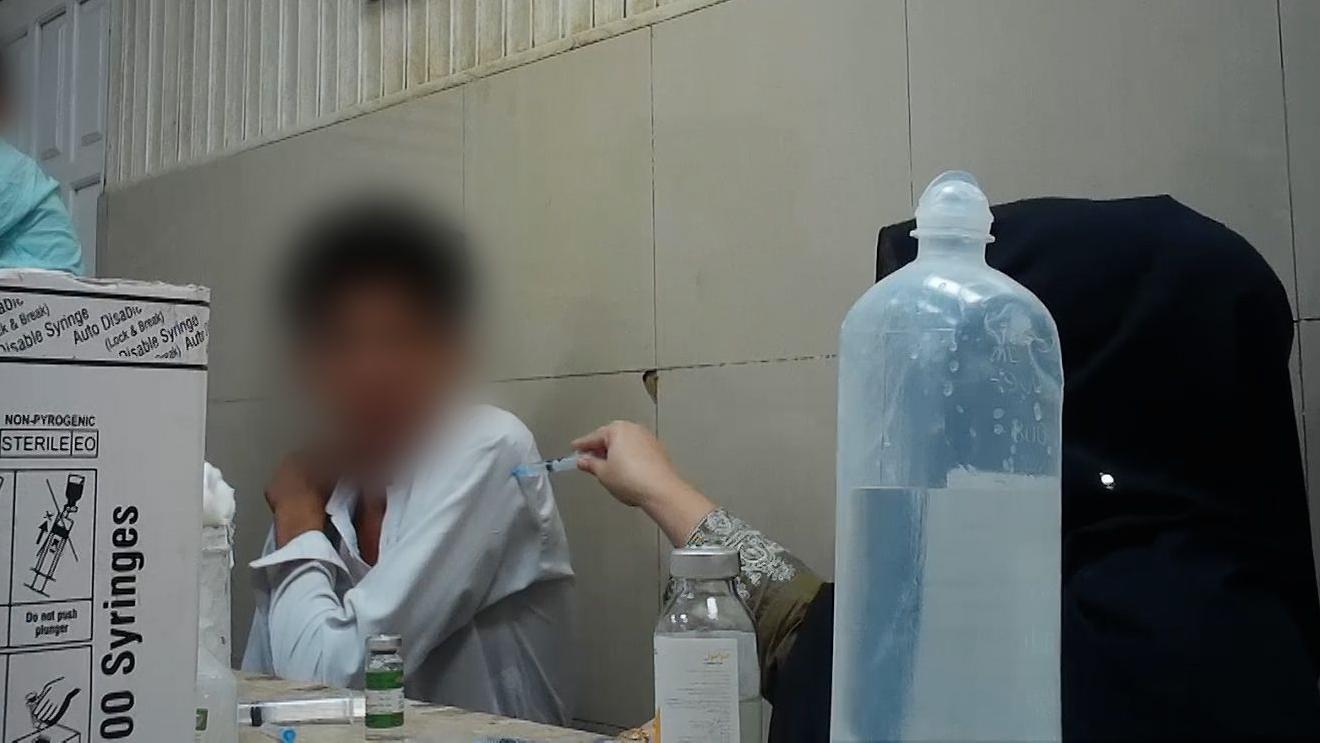 Una escena en un hospital. Una mujer, cuyo rostro no se ve, est&aacute; poniendo una inyecci&oacute;n a un paciente, cuyo rostro tambi&eacute;n est&aacute; borroso. Le est&aacute; poniendo la inyecci&oacute;n sin guantes y clav&aacute;ndole la aguja a trav&eacute;s de la manga de la camisa.