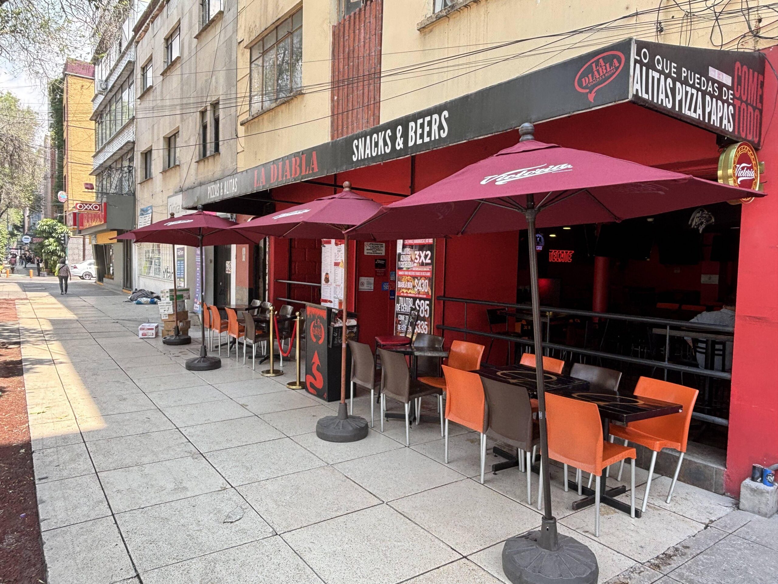 Terraza de restaurante en la acera con mesas y sillas bajo sombrillas rojas, frente a una fachada urbana con letreros.
