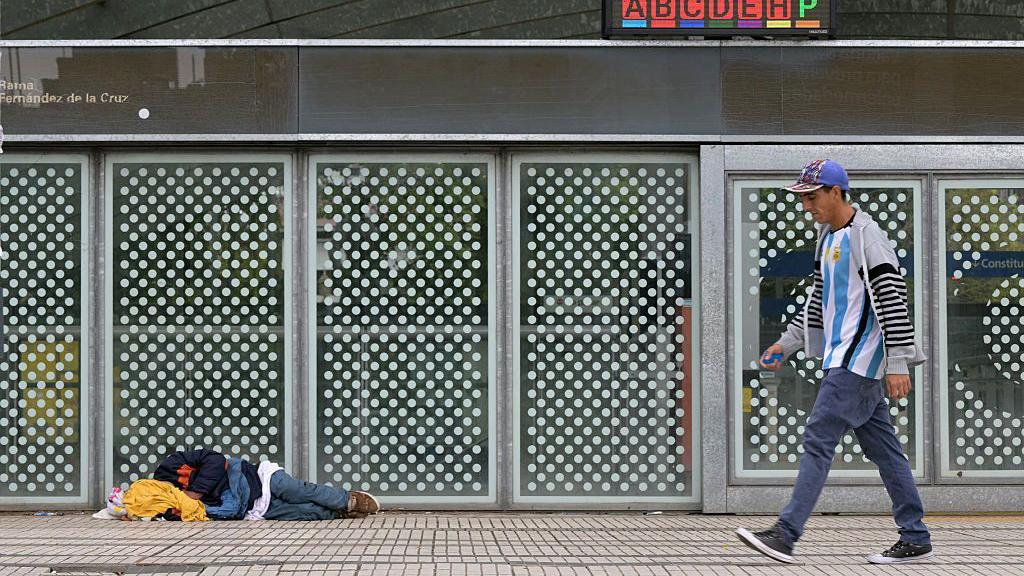 Un hombre pasa frente a la estaci&oacute;n de metro Constituci&oacute;n, en Buenos Aires, mientras una persona duerme en el suelo.