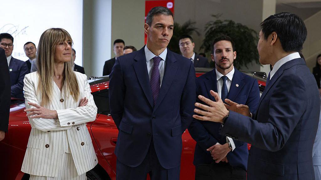 Pedro S&aacute;nchez, en medio, vestido con traje y corbata conversa con un funcionario chino (a la derecha) durante un viaje oficial. Bego&ntilde;a est&aacute; de pie a la izquierda con los brazos cruzados. Es una mujer rubia, delgada y viste un chaleco color blanco. En el fondo se ven varios hombres tambi&eacute;n vestidos de traje y un auto rojo. 