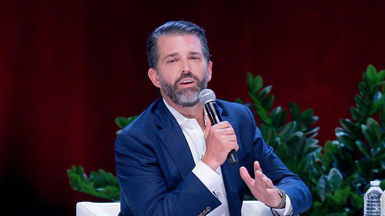 Donald Trump Jr, hijo de presidente, con un micr&oacute;fono en mano, de chaqueta azul y camisa blanca, habla en una conferencia en Sugar Land, Texas. 23 de octubre 2025.