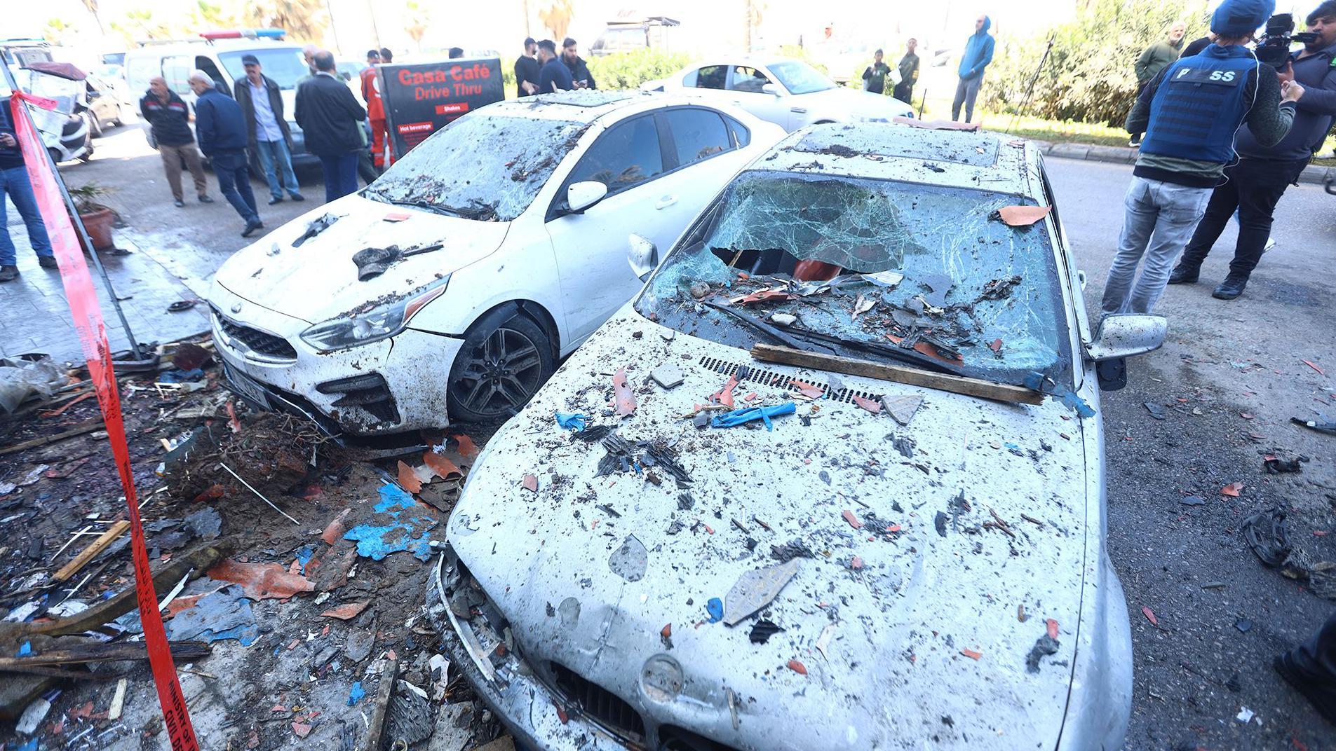 En la imagen aparecen dos autom&oacute;viles da&ntilde;ados y cubiertos de escombros, con multitudes de personas en la calle de fondo y ruinas tras un ataque con misiles israel&iacute;es en la carretera costera de Sid&oacute;n este mi&eacute;rcoles.