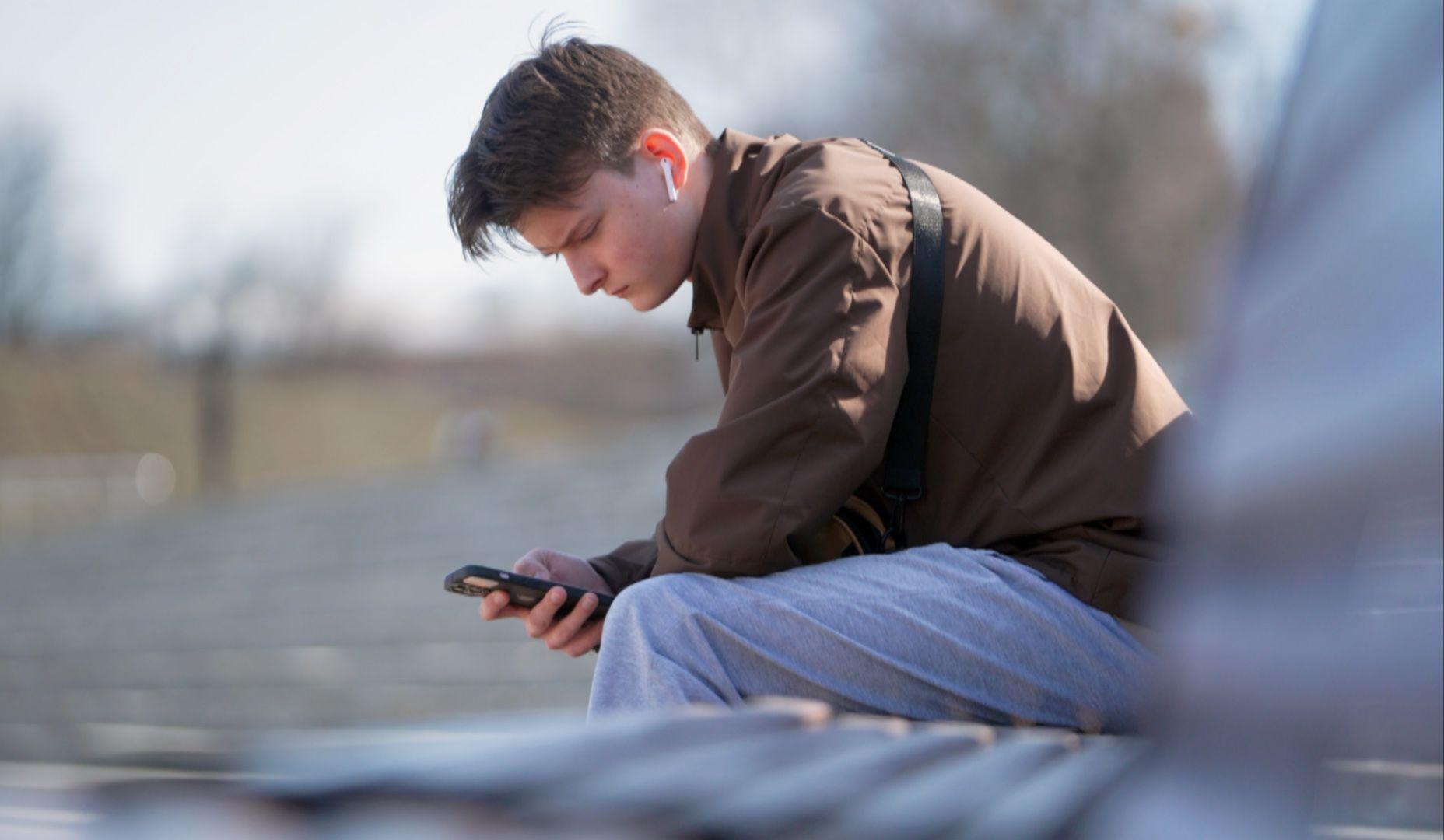 Un joven usa su tel&eacute;fono m&oacute;vil mientras est&aacute; sentado en un banco de un parque en un d&iacute;a despejado en Vladimir.