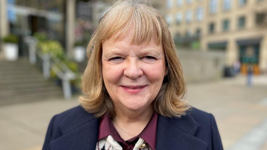Una mujer que sonr&iacute;e a la c&aacute;mara, tiene el pelo rubio y lleva una chaqueta azul oscuro sobre una blusa burdeos y blanca