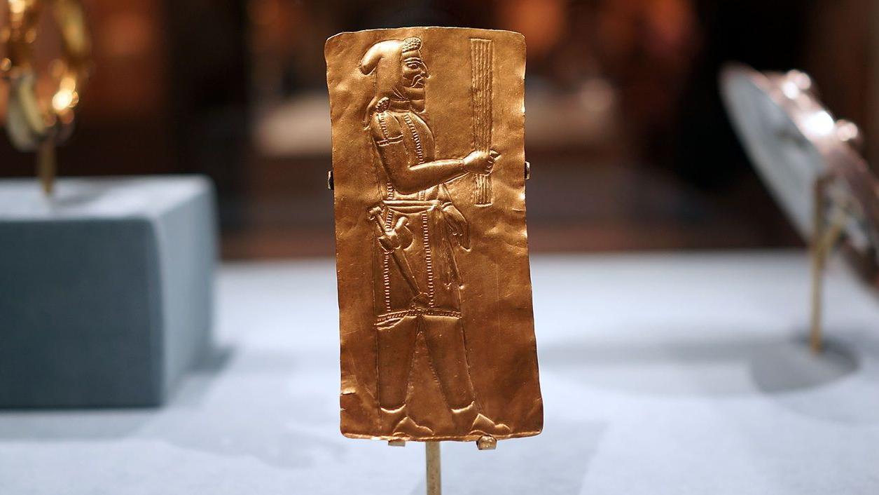 Una placa dorada en la que se ve un hombre de perfil llevando un objeto largo en la mano. Viste un gorro, una t&uacute;nica corta y pantalones. Lleva una especie de espada en la cintura.
