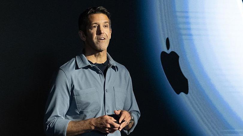 John Termus, con camisa gris, frente a una pantalla que proyecta el logotipo de Apple, durante la presentaci&oacute;n de un producto.