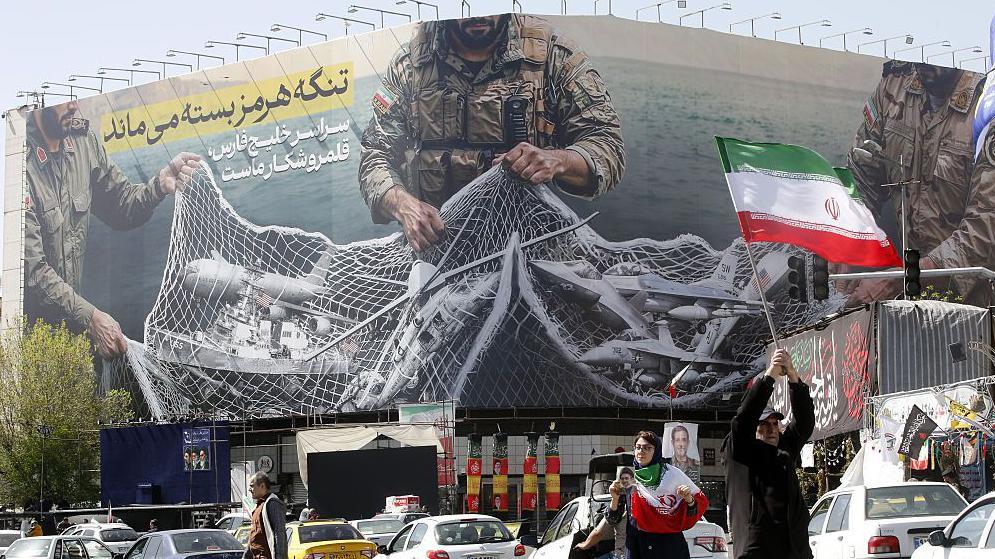 Imagen de una gran pancarta colgada en Teher&aacute;n, en la que se lee: &laquo;El estrecho de Ormuz permanecer&aacute; cerrado...&raquo; &laquo;Todo el golfo P&eacute;rsico es nuestro coto de caza&raquo;; la imagen muestra a soldados sosteniendo una red de pesca en la que han quedado atrapados buques cisterna y aeronaves militares. En primer plano aparecen autom&oacute;viles y personas que sostienen o ondean banderas iran&iacute;es.