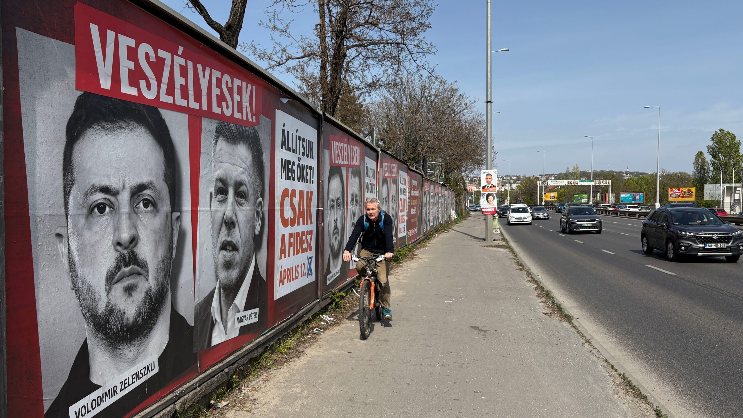 Un hombre pasa en bicicleta junto a un muro en una autopista de Hungr&iacute;a, empapelado con retratos de estilo cine negro de Volodymyr Zelensky y P&eacute;ter Magyar.