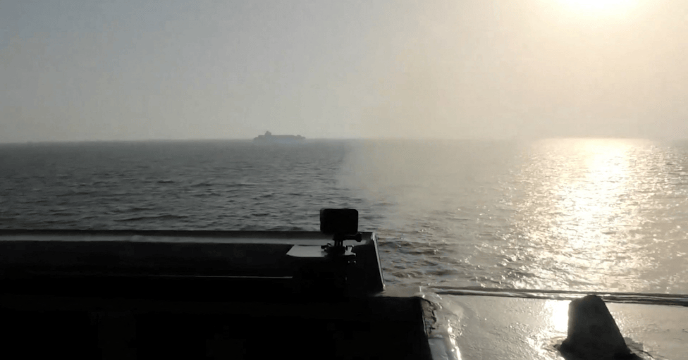 Un barco de carga en el horizonte visto desde la cubierta de otro barco del que sale humo. 