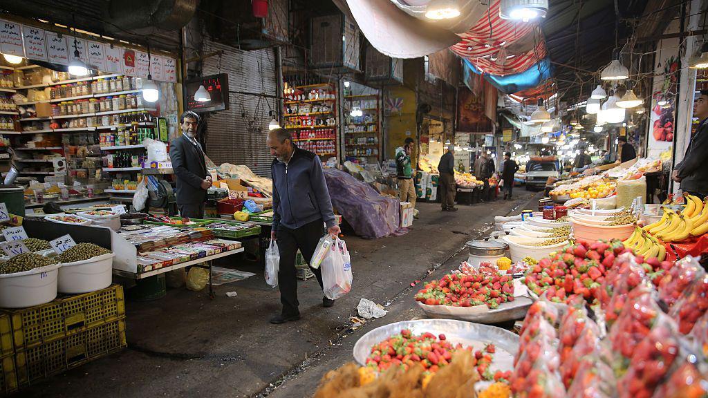 Un hombre carga bolsas en ambas manos mientras camina dentro de un mercado de alimentos.