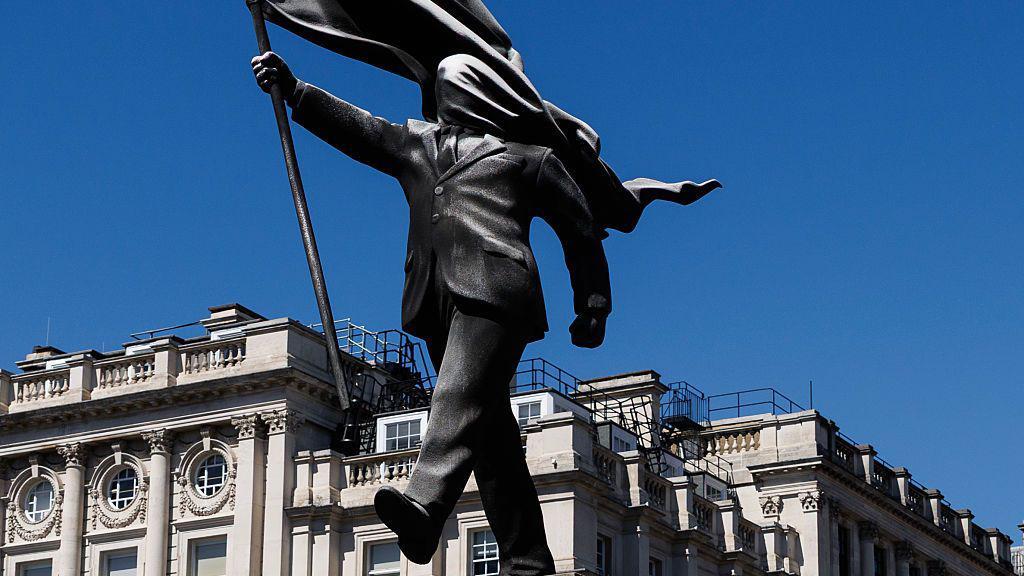 Una estatua que representa a una persona descendiendo de un pedestal con el rostro cubierto por una bandera ondeante en Waterloo Place, el 30 de abril de 2026, en Londres, Inglaterra.