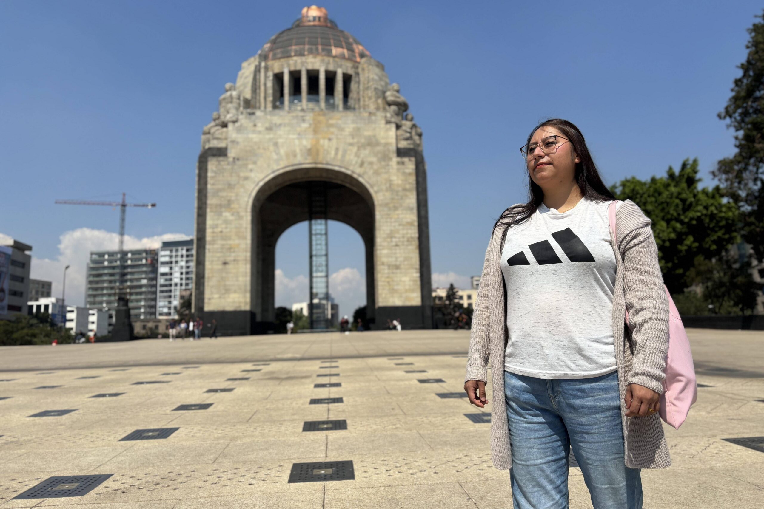 Mujer de pie en una plaza amplia frente al Monumento a la Revoluci&oacute;n, con edificios urbanos al fondo y cielo despejado.