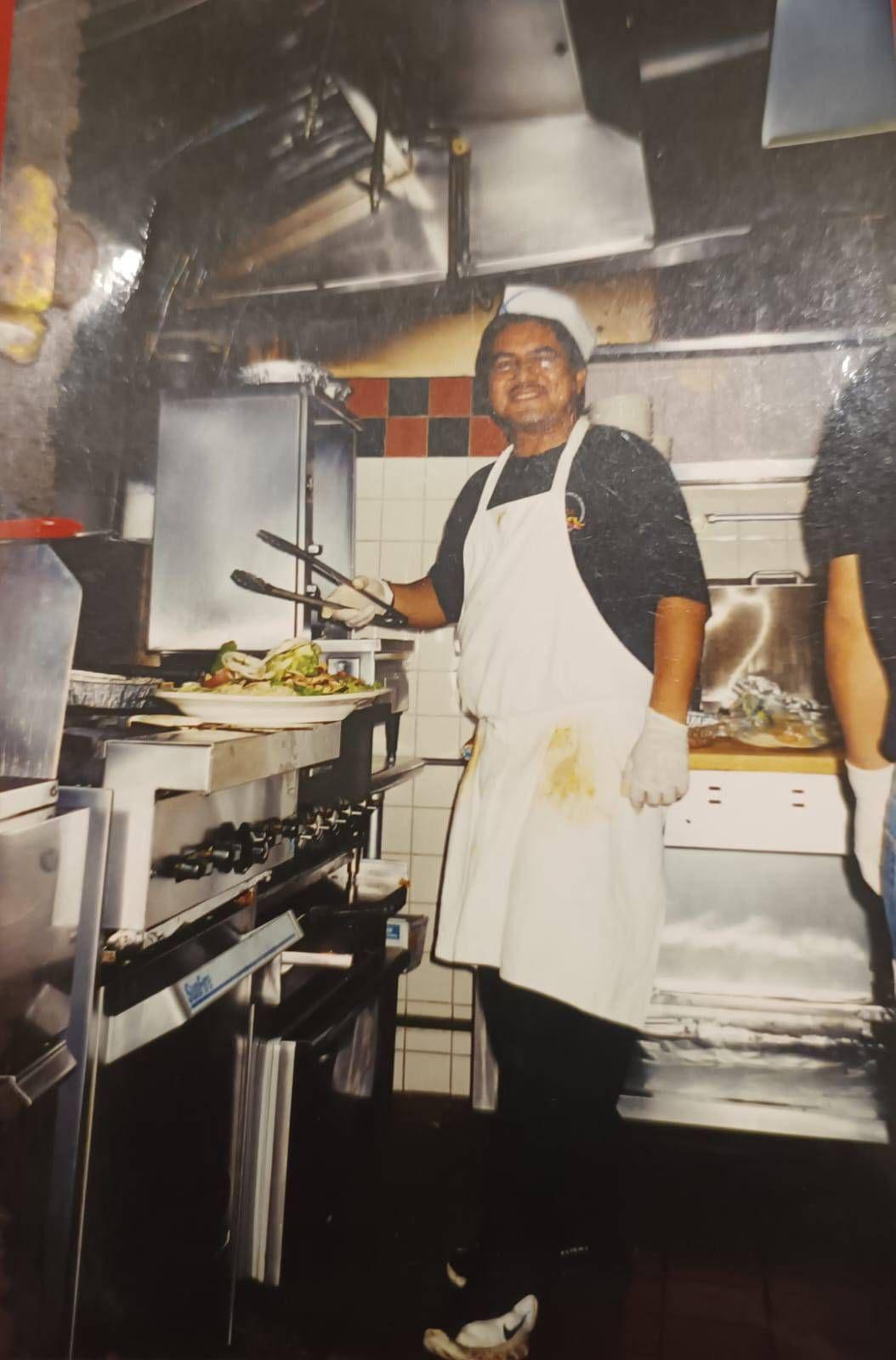 Hombre con delantal trabajando en una cocina industrial, usando pinzas frente a una parrilla con comida, rodeado de equipos de metal.