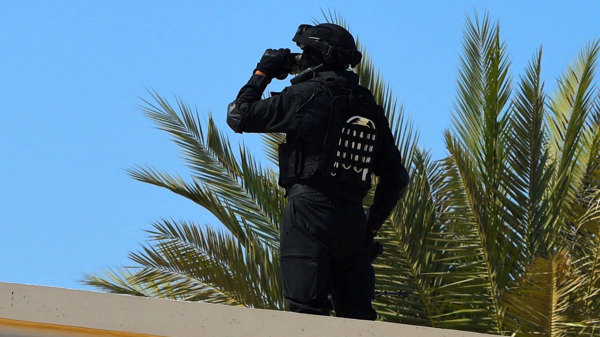 Un guardia de seguridad bahrein&iacute; se encuentra sobre una azotea junto a una palmera en un d&iacute;a soleado. Se lleva la mano al rostro y viste un uniforme negro con casco negro.