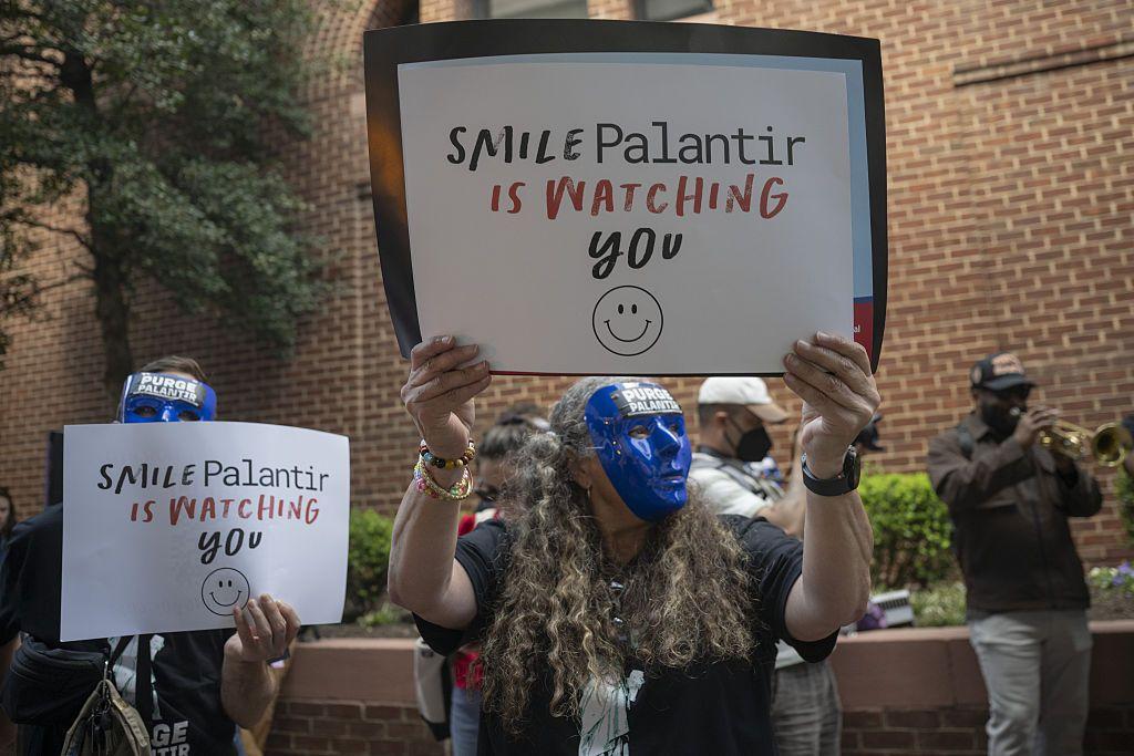 Manifestantes con m&aacute;scaras azules sostienen letreros que leen "Sonr&iacute;e, Palantir te est&aacute; mirando".