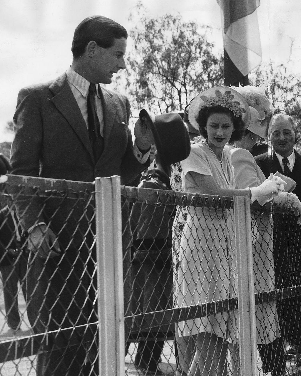 Un hombre alto con traje formal sostiene un sombrero en su mano y mira atentamente a una joven mujer vestida de blanco con un sombrero con flores. Ambos est&aacute;n al aire libre detr&aacute;s de una verja 