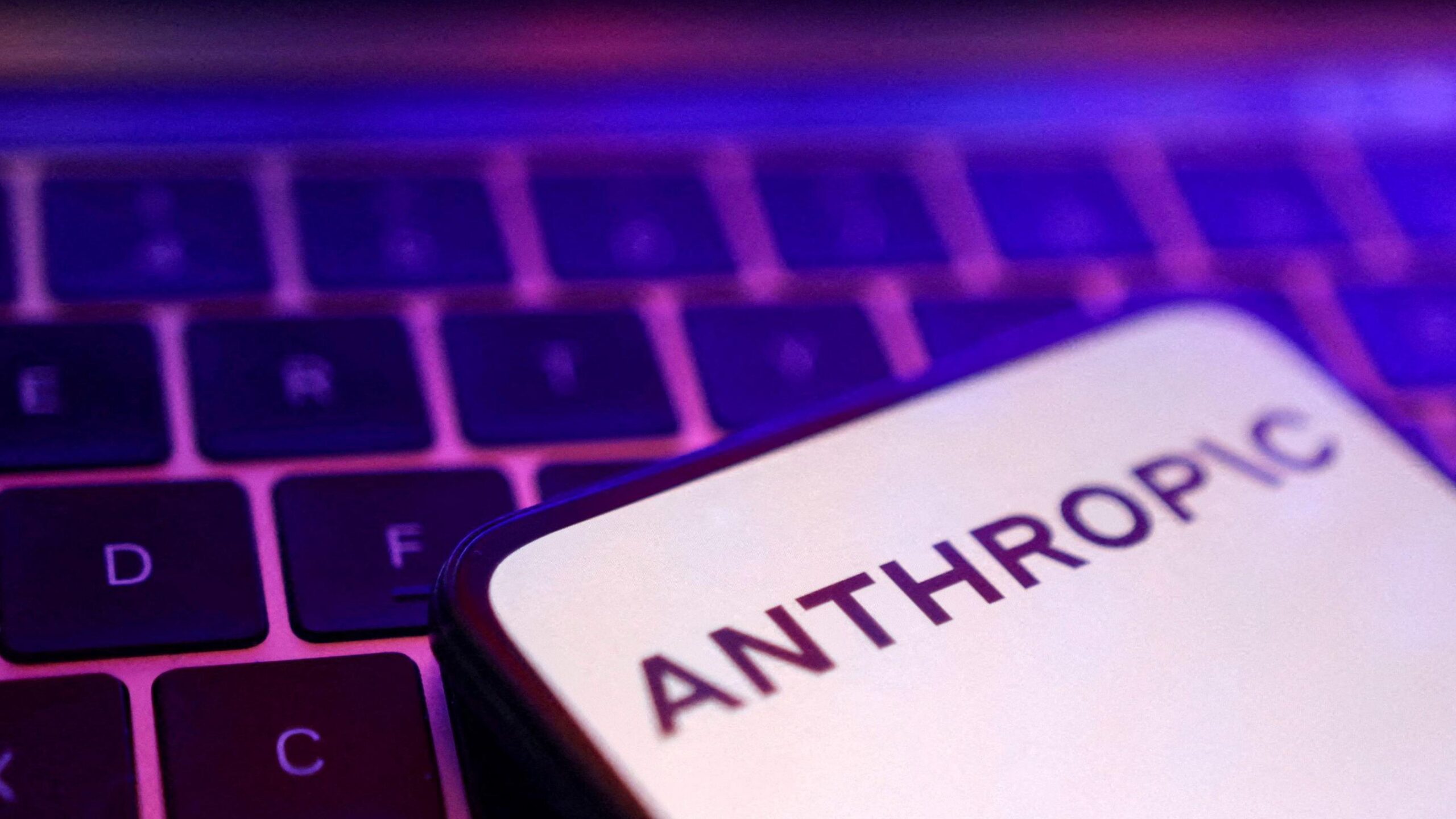 Una pantalla de smartphone que muestra el logotipo de Anthropic en letras negras sobre un fondo completamente blanco, colocada sobre un teclado de port&aacute;til iluminado en rosa y morado.