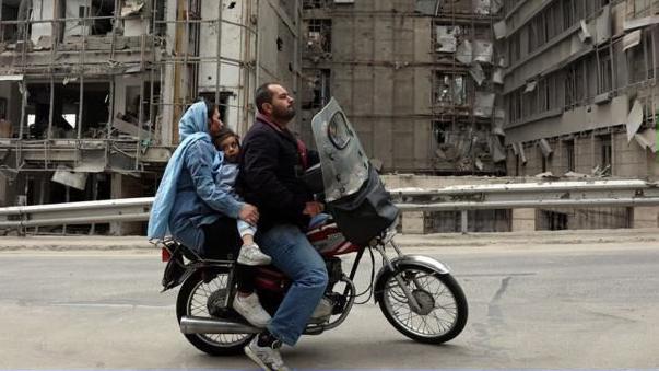 Una pareja viajando en motocicleta con un ni&ntilde;o en medio. Detr&aacute;s hay esdificios destruidos.