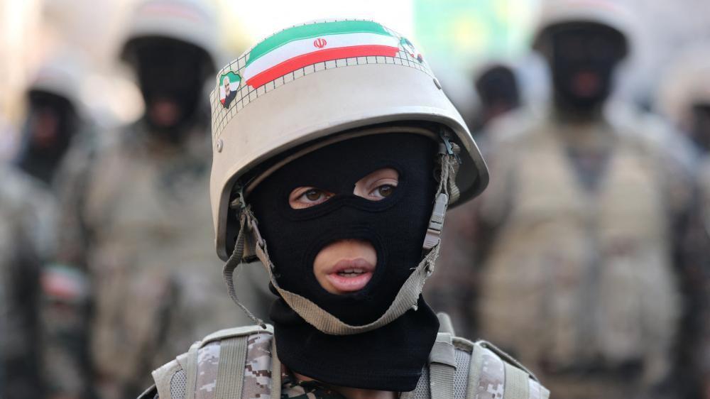 Un joven que lleva un pasamonta&ntilde;as, uniforme de fatiga y un casco con una bandera iran&iacute; mira fuera de c&aacute;mara, con figuras militares borrosas en el fondo.