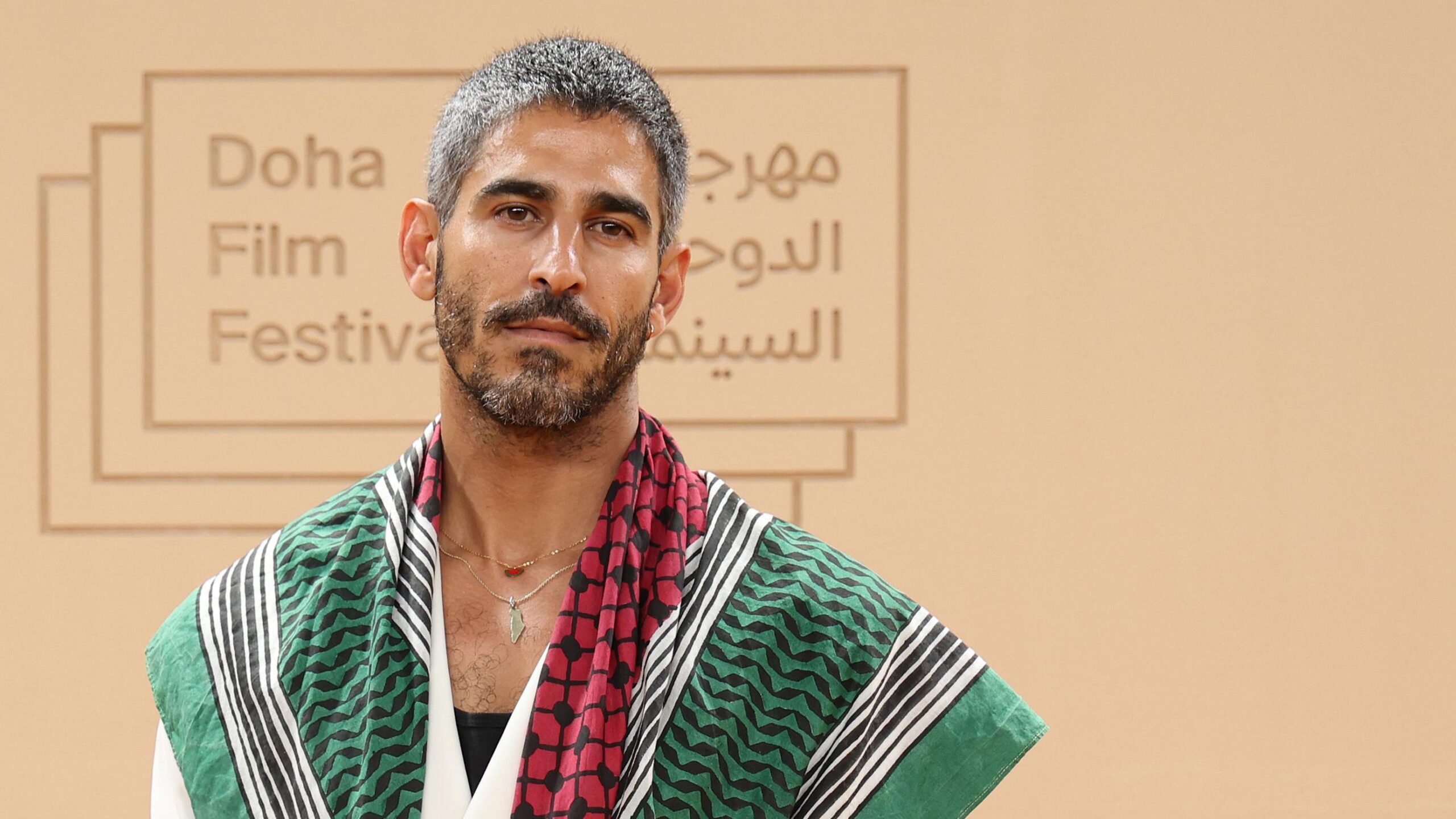 Una foto del periodista kuwait&iacute;‑estadounidense Ahmed Shihab‑Eldin, con el logotipo del Festival de Cine de Doha 2025 visible en el fondo. Tiene el cabello gris y una barba negra y gris, y viste una prenda con un patr&oacute;n verde, rojo y blanco, adem&aacute;s de un collar.