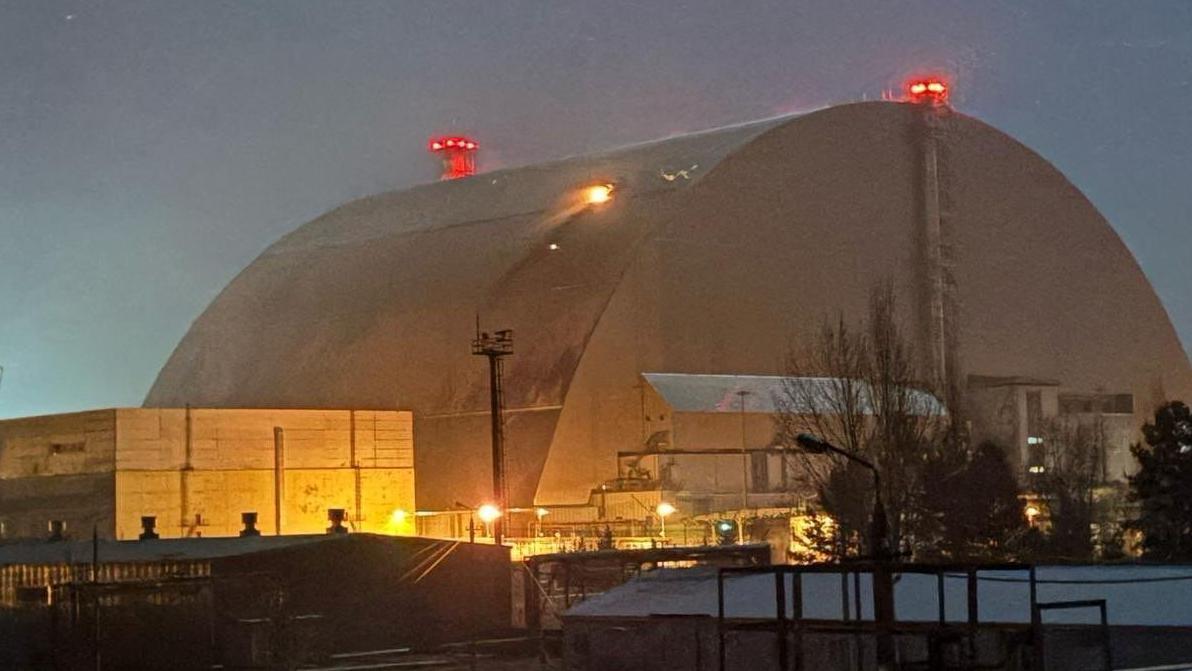 El gran escudo met&aacute;lico curvado que cubre el reactor cuatro, fotografiado con poca luz, con llamas saliendo de un agujero en uno de sus lados, donde fue alcanzado por un dron el 14 de febrero de 2025. Se observan luces rojas en la parte superior de la c&uacute;pula y luces amarillas alrededor de los edificios m&aacute;s bajos situados cerca de ella.