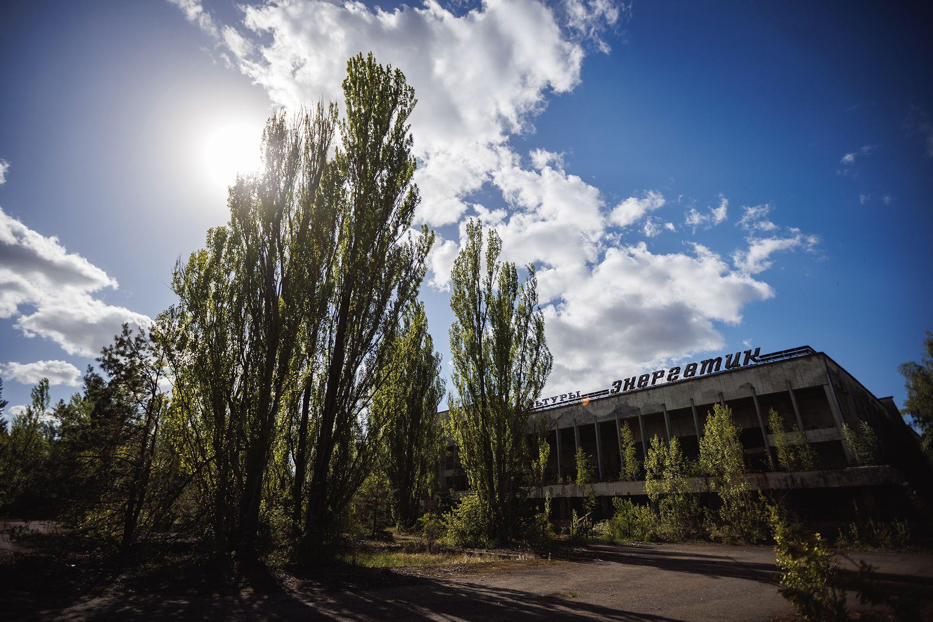 Vista exterior del abandonado Palacio de la Cultura Enerhetyk en la plaza central de Pripyat, &oacute;blast de Kiev, Ucrania, el 1 de mayo de 2025. Grandes &aacute;rboles verdes crecen frente a &eacute;l y se ve contra un cielo azul con nubes blancas.