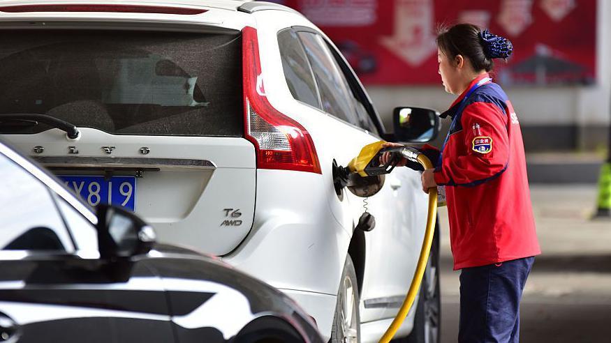 Una empleada de una gasolinera junto a un coche blanco.