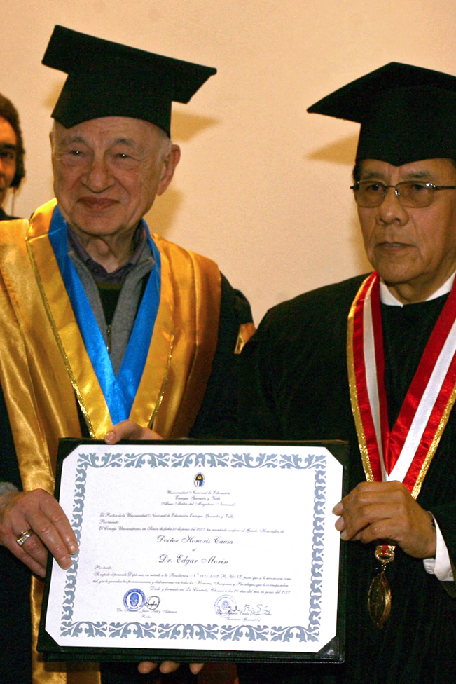 Edgar Morin con la toga y birrete y una medalla de cinta azul y amarilla sostiene el t&iacute;tulo junto al rector Juan Tutuy Aspauza, quien tambi&eacute;n llevaba toga y birrete y una medalla con cinta roja y blanca. 
