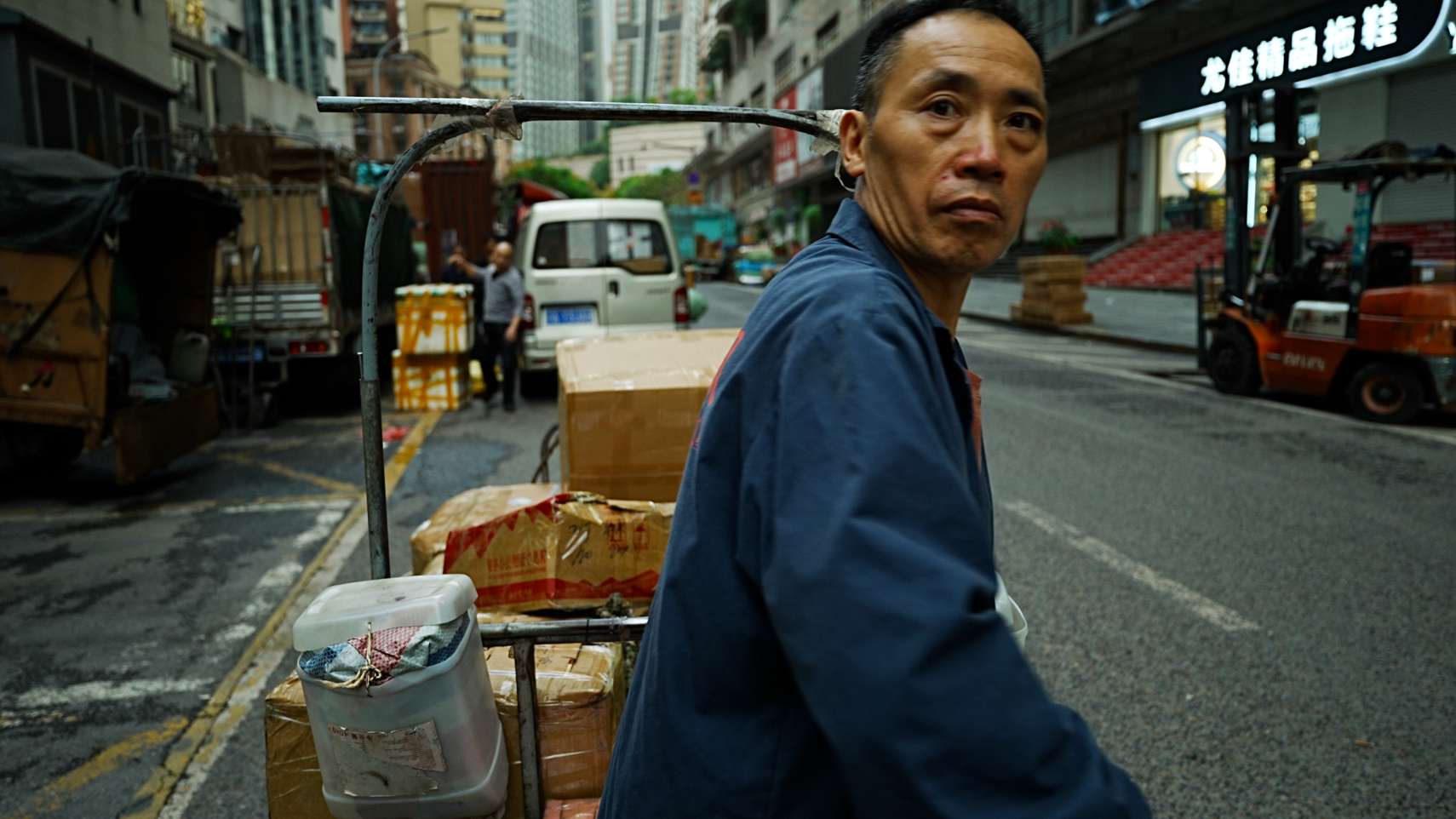 Un trabajador de Chongqing vestido con un mono azul tirando de un carro en una calle de rascacielos.