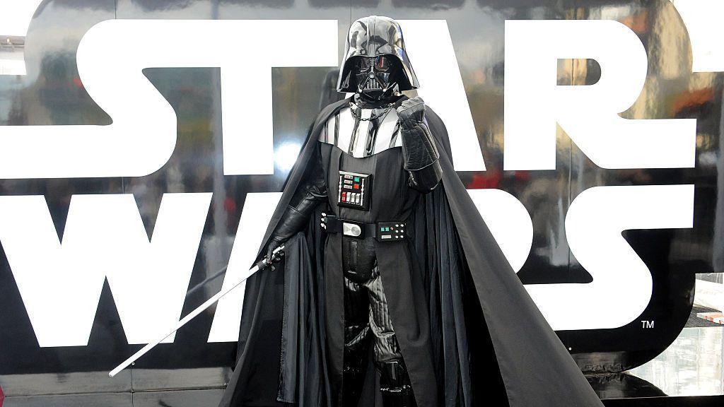 imagen de una persona con un traje de darth vader. tiene el pu&ntilde;o cerrado y levantado hacia el frente y en la otra mano sostiene un sable de luz. 