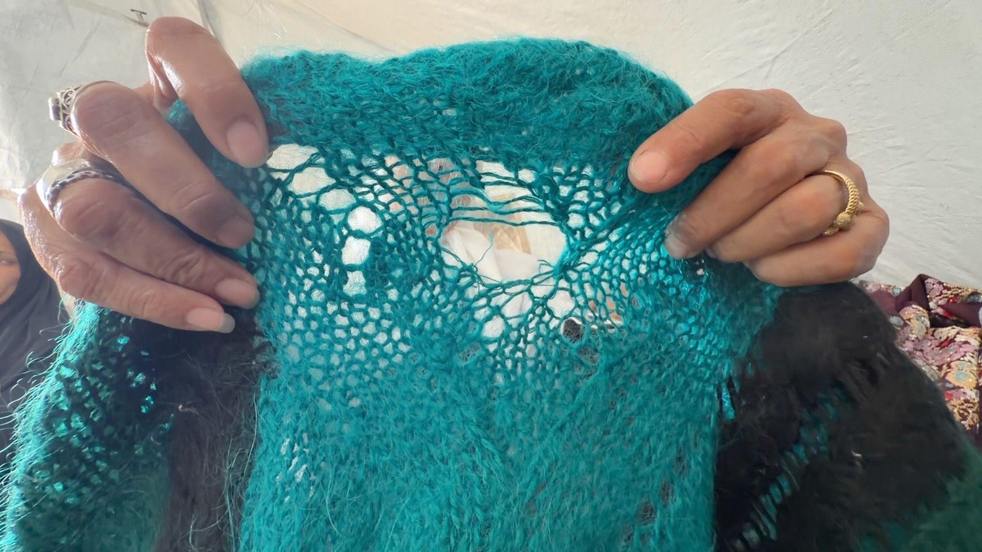 Los dedos con anillos de Amani Abu Salmiya sostienen una prenda de punto azul con un agujero en el centro.