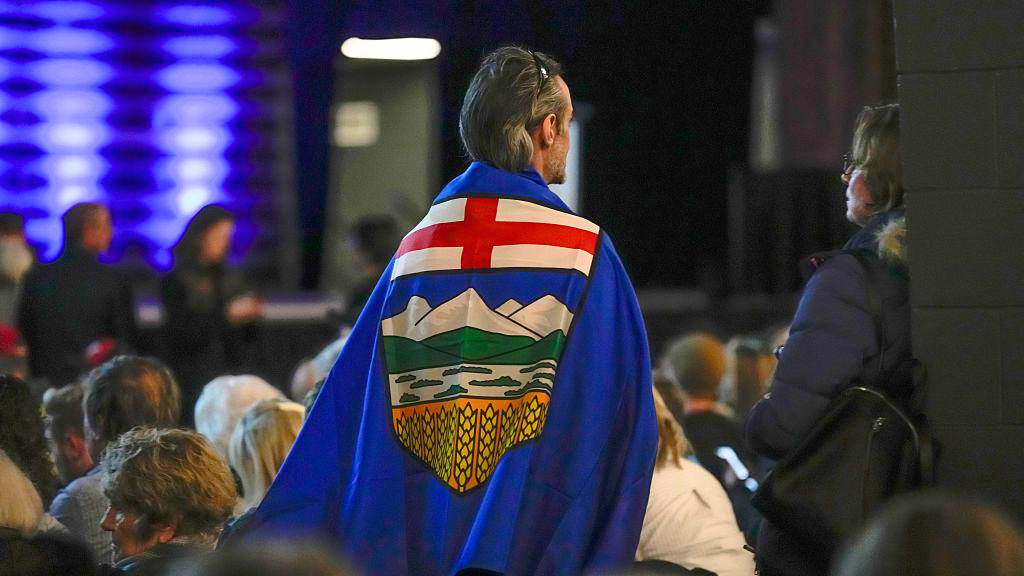 Un adepto lleva una bandera de Alberta colgada a la espalda mientras se pone de pie durante un evento en el Ayuntamiento de Independencia de Alberta en Calgary, Alberta, Canad&aacute;