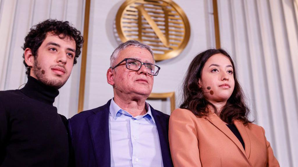 
Taghi Rahmani, Ali Rahmani y Kiana Rahmani posan para una fotograf&iacute;a durante una conferencia de prensa en el Instituto Nobel. El logotipo dorado del Nobel se ve en el fondo. Los tres llevan peque&ntilde;os tocados con micr&oacute;fono.