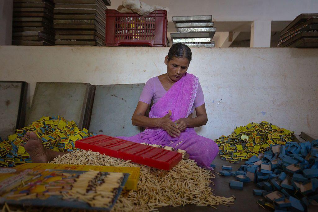Una mujer tamil vestida con un sari rosa est&aacute; sentada en el suelo, empaquetando cientos de cajas de cerillas 