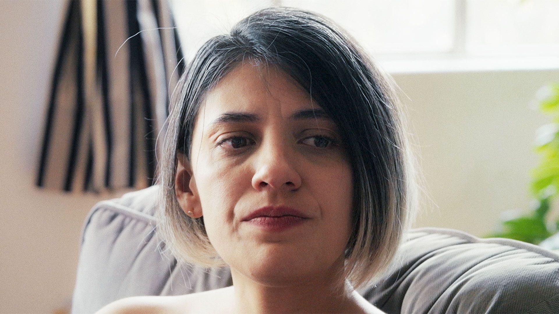 Alex lleva el cabello corto, tiene los ojos oscuros, lleva pintalabios rojo, est&aacute; sentada en un sof&aacute; y tiene un aspecto apesadumbrado.