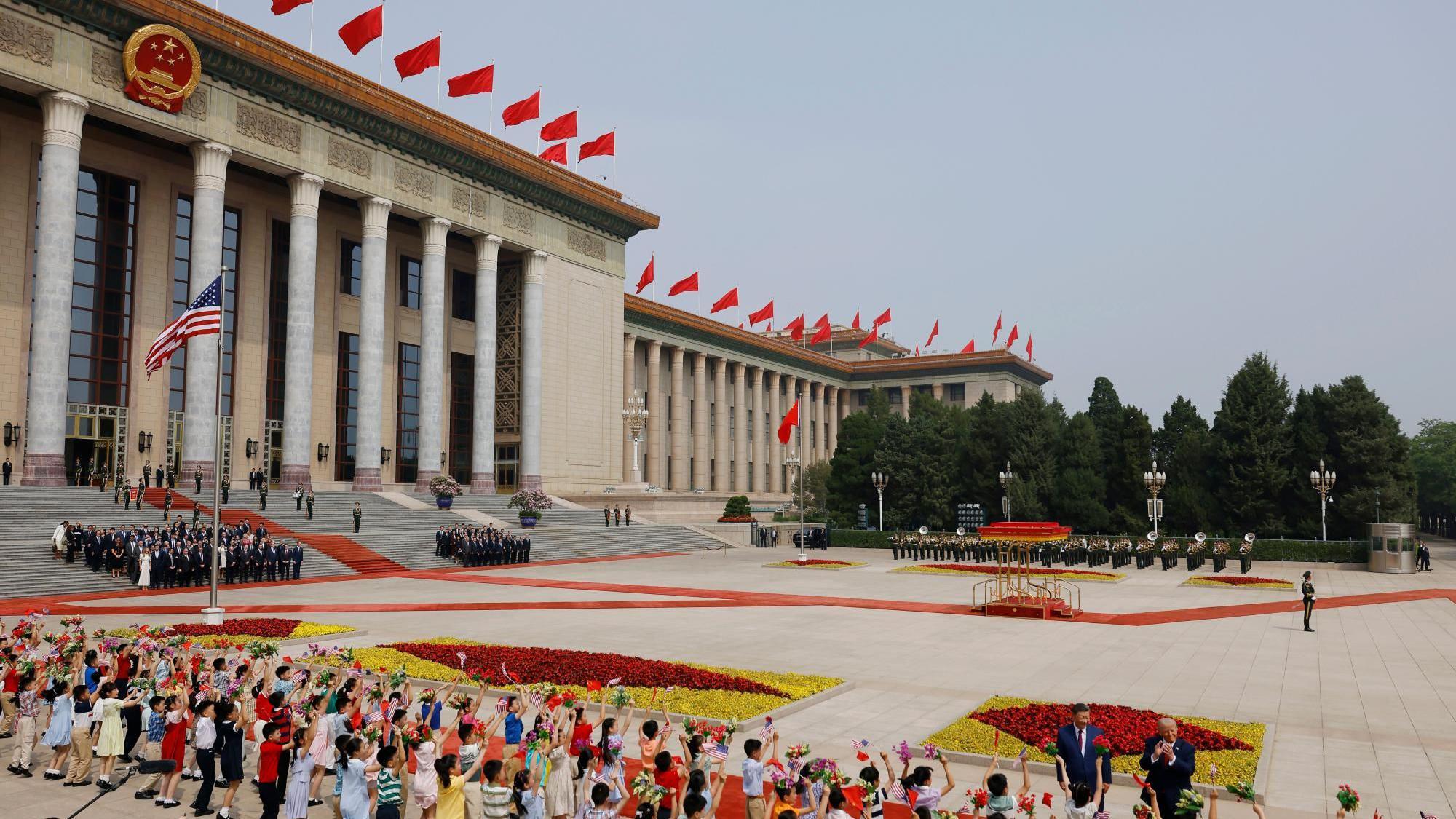 Una vista amplia de una plaza pavimentada con banderas chinas y estadounidenses izadas, y un largo edificio con columnas a la izquierda que luce banderas chinas en su tejado; en primer plano, se observa a los presidentes Xi y Trump saludando a la multitud.