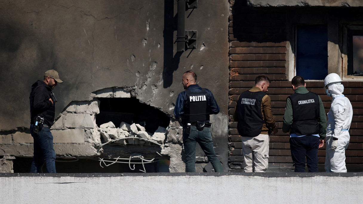 Cinco miembros de las fuerzas del orden de Rumania inspeccionan el lugar donde se produjo el choque del dron. Cuatro de ellos est&aacute;n vestidos de civil y llevan chalecos negros. Uno tiene un traje blanco y un casco. En la pared se ve un agujero