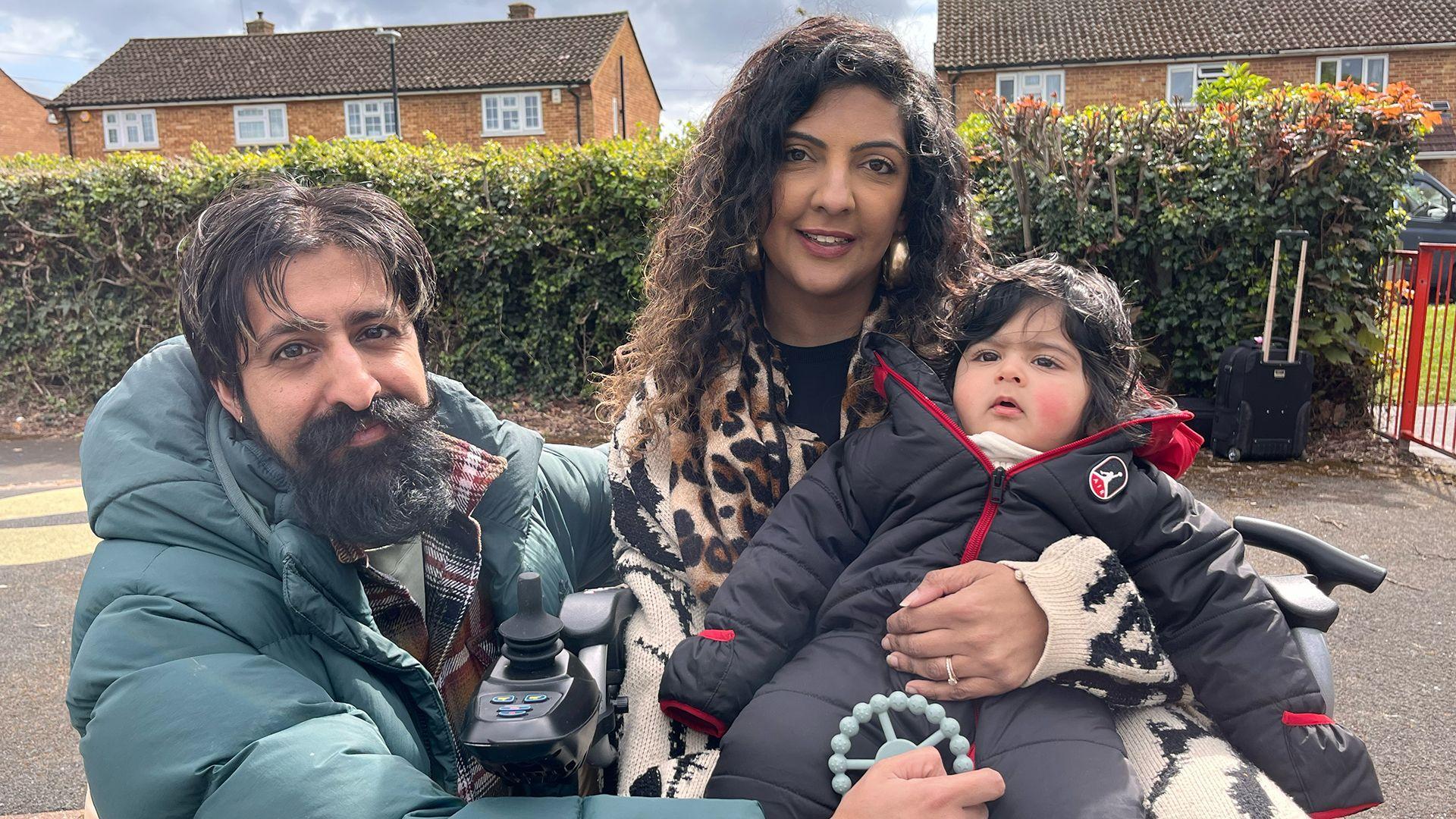Un hombre y una mujer sostienen a un ni&ntilde;o peque&ntilde;o. El hombre tiene el pelo corto y oscuro, barba y lleva una chaqueta acolchada de color verde gris&aacute;ceo. La mujer tiene el pelo largo, oscuro y ondulado, y lleva una bufanda con estampado de leopardo. El ni&ntilde;o peque&ntilde;o tiene el pelo oscuro y lleva un mono acolchado gris oscuro con cremallera.