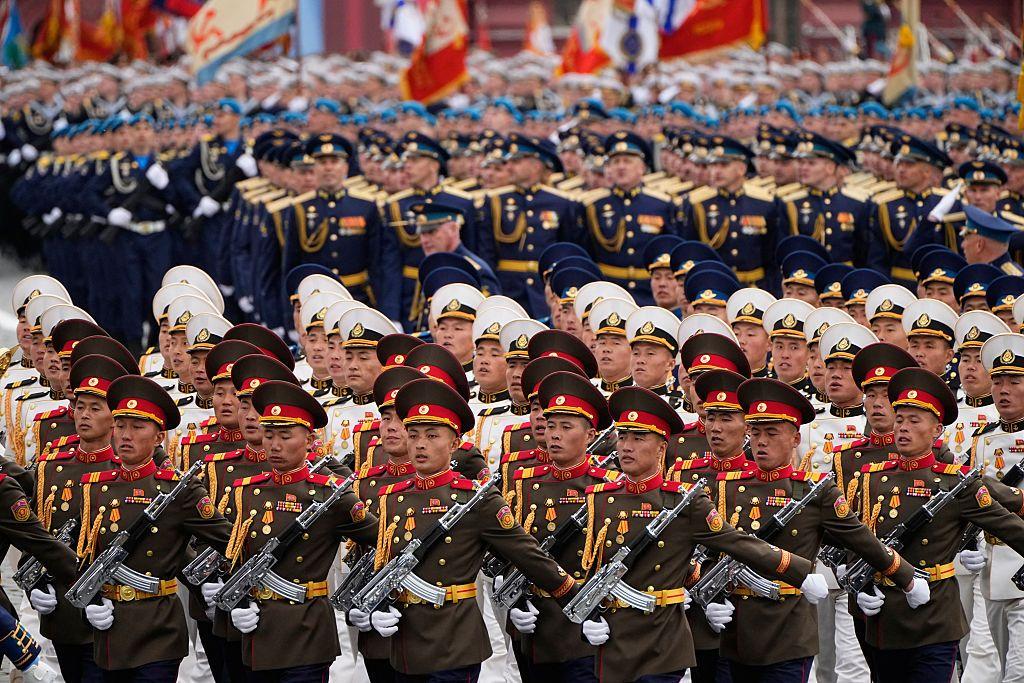 Soldados norcoreanos desfilan en filas portando armas. Los soldados de las primeras filas visten uniformes de color caqui con galones dorados y rojos. M&aacute;s atr&aacute;s se encuentran otros soldados norcoreanos con uniformes blancos con galones dorados y negros. Detr&aacute;s de ellos hay filas de soldados rusos.