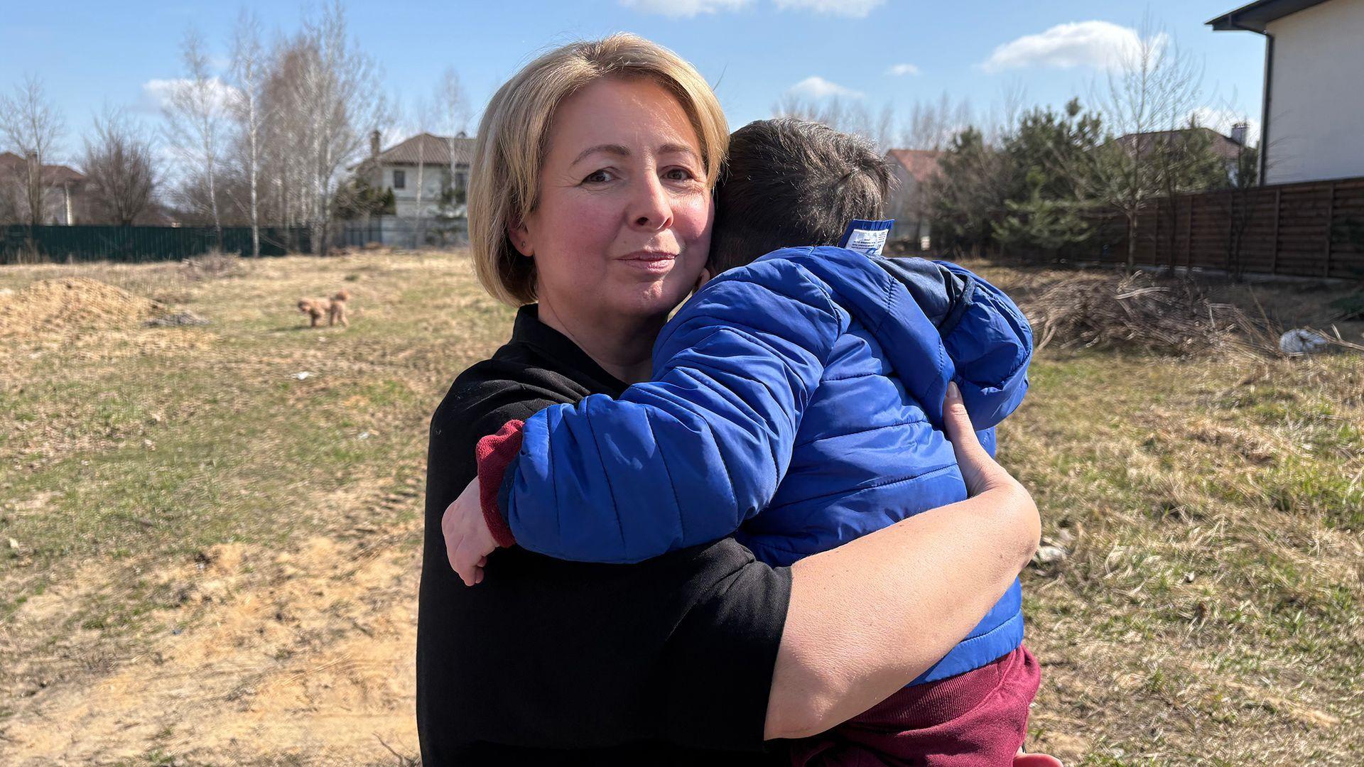 Una mujer sostiene a un ni&ntilde;o peque&ntilde;o en brazos. El ni&ntilde;o est&aacute; de espaldas a la c&aacute;mara, por lo que no se ve su rostro. Tiene el pelo oscuro y lleva una chaqueta azul y pantalones rojos. La mujer mira a la c&aacute;mara; tiene el pelo corto, liso y rubio, y lleva una blusa negra. Est&aacute;n al aire libre y se puede ver hierba alta y algunas casas detr&aacute;s.