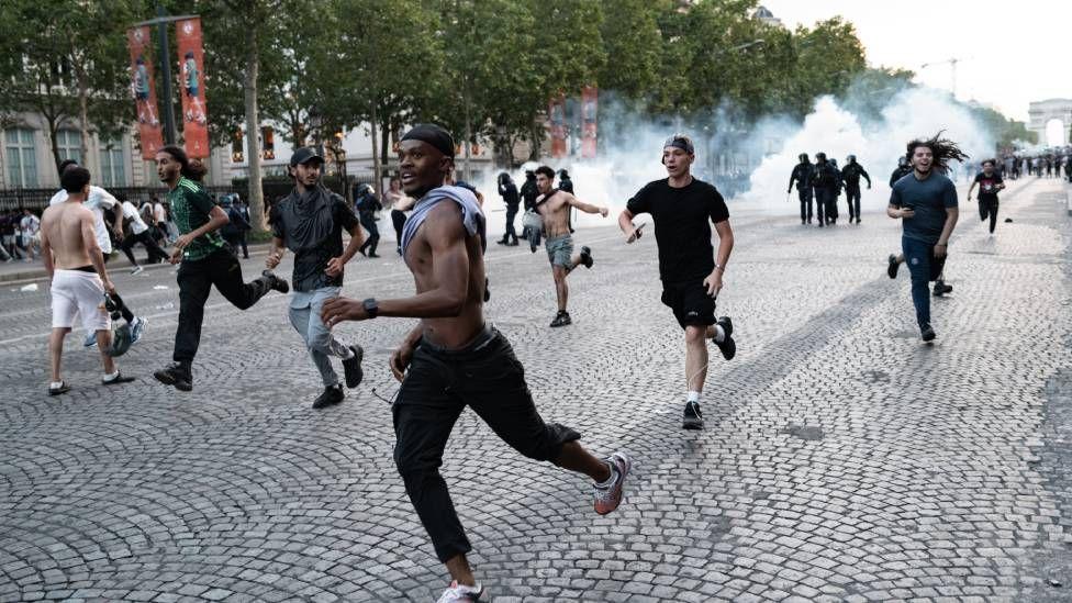 Varios j&oacute;venes corren por una calle, al fondo se ver humo
