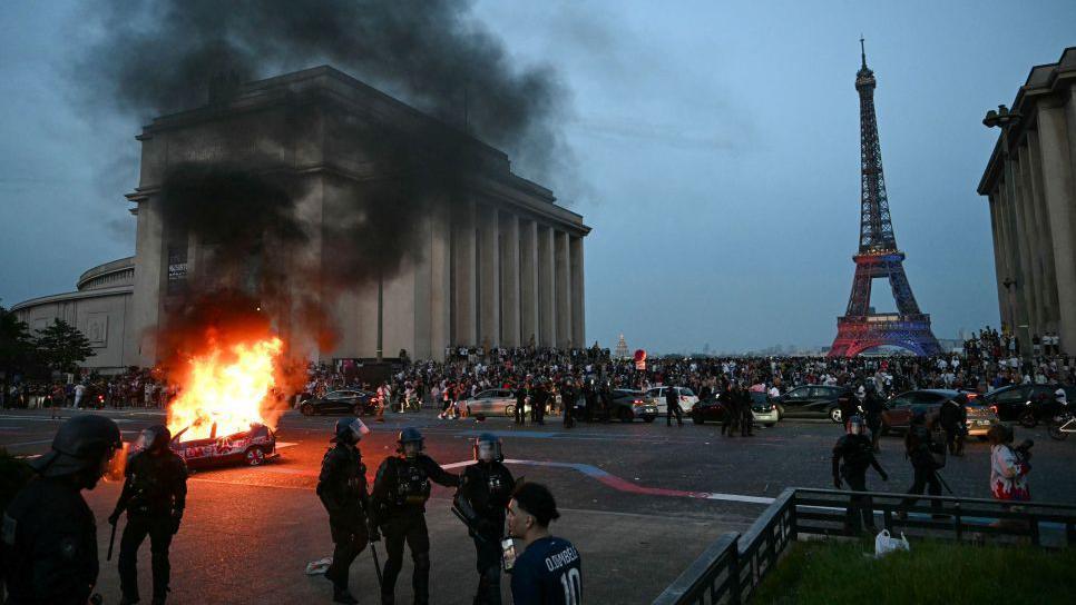 A la izquierda se ve un veh&iacute;culo en llamas. Muy cerca est&aacute; un grupo de polic&iacute;as. Al fondo se ven veh&iacute;culos de la polic&iacute;a, una concentraci&oacute;n de personas y la Torre Eiffel