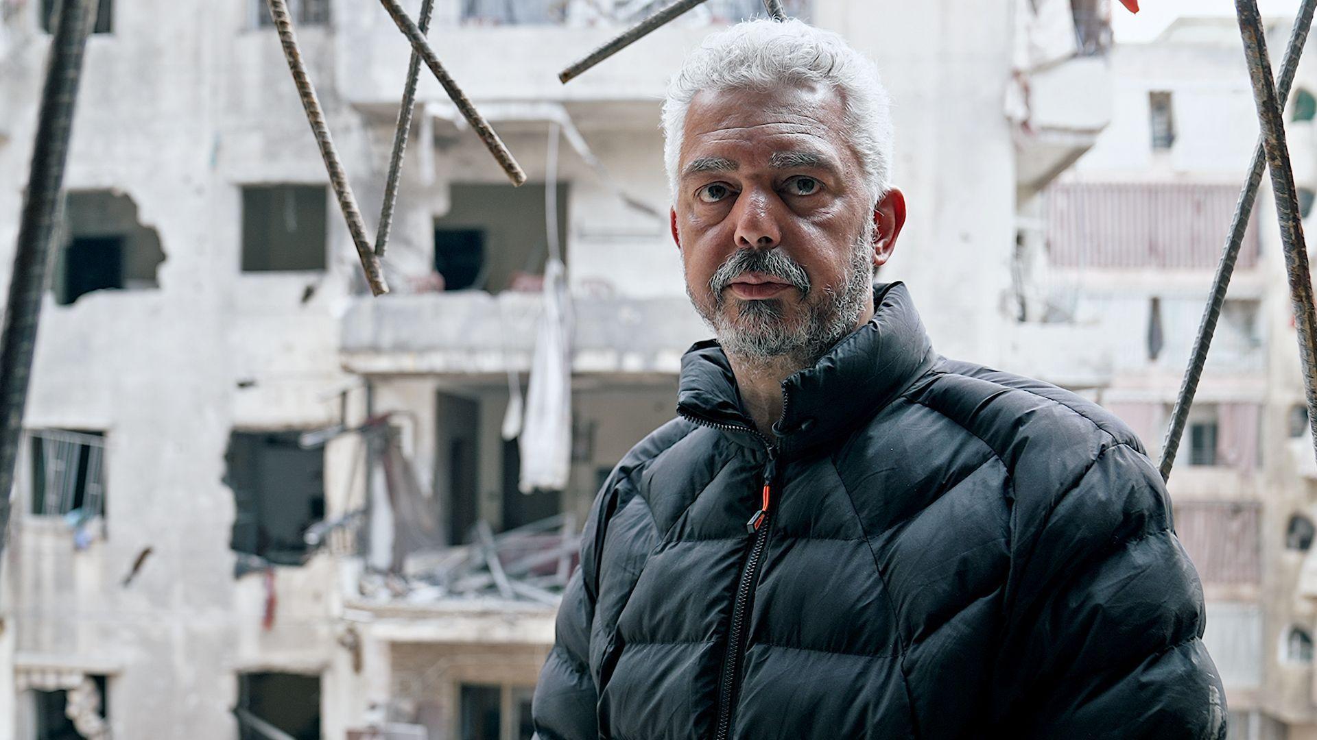 Mohammed, un hombre en sus 40, con pelo y barba canosos, est&aacute; parado frente a un edificio en ruinas.