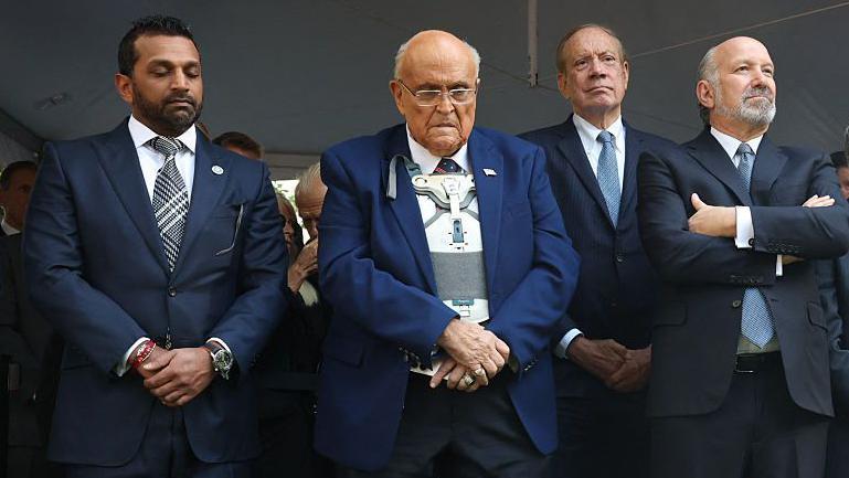 Giuliani, en el centro, con una chaqueta azul sobre un soporte m&eacute;dico blanco, cruza las manos y mira hacia abajo. Est&aacute; flanqueado por Kash Patel y Howard Lutnick.
