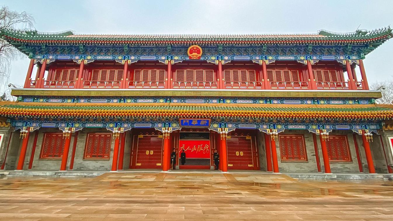 Un edificio tradicional chino ornamentado, con pilares rojos y grandes portones rojos en el centro; varios guardias permanecen de pie en la entrada.
